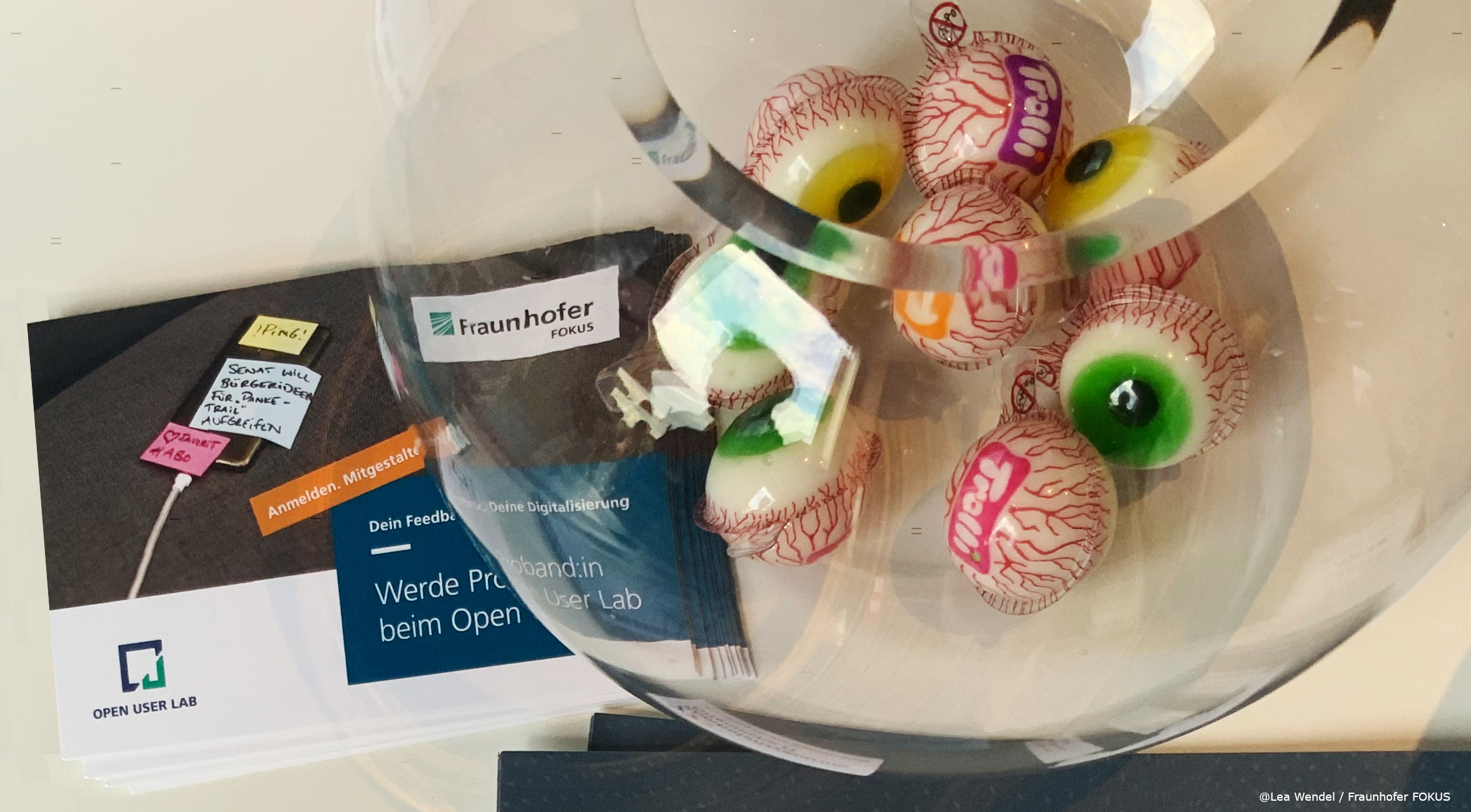 Auf einem Tisch liegen Flyer mit dem Aufdruck "Werde Proband:in beim Open User Lab", darüber steht eine runde Vase mit Trolli Süßigkeiten Augen